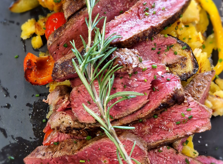 Several slices of pink roasted beef steak on a black plate, garnished with a sprig of rosemary, served with colourful pieces of bell pepper and mashed potatoes.