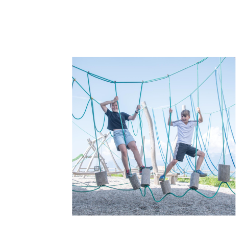 Two children on wooden blocks in the rope climbing park near a small playground under blue skies.