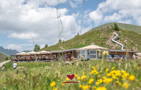 A mountain restaurant surrounded by a small playground in the middle of green countryside, with mountains and blue skies in the background.